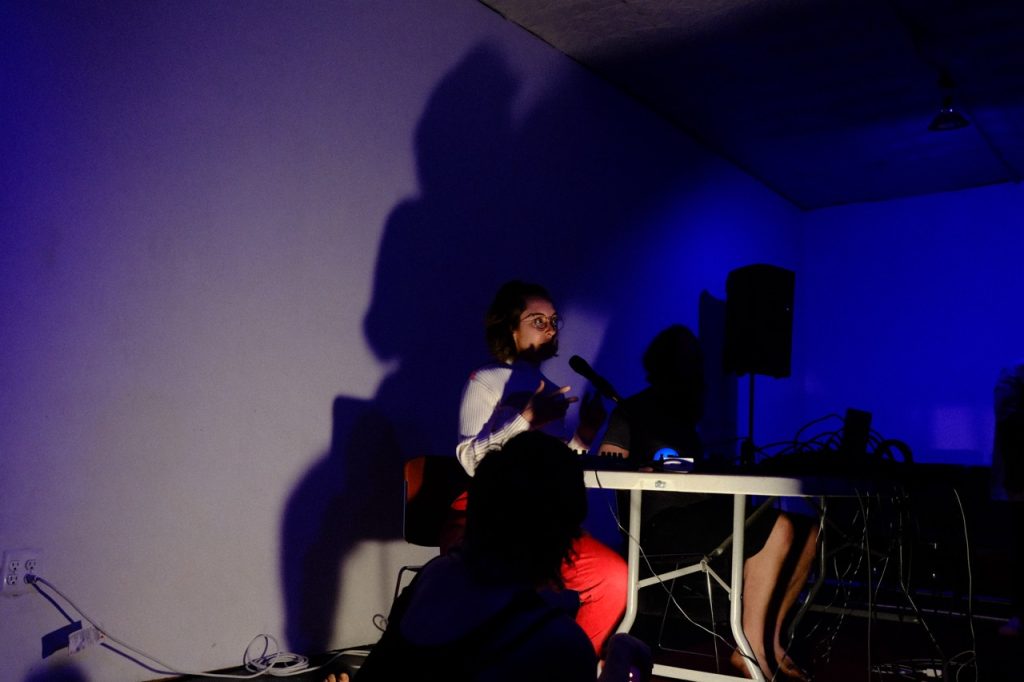 Image: Paige Naylor performs at EXP/VOX. She sits at a table with a synthesizer and microphone positioned in front of her. Naylor's shadow is cast on the white wall behind her. Photo by Michael Wellvang. Image courtesy of Anna Johnson.