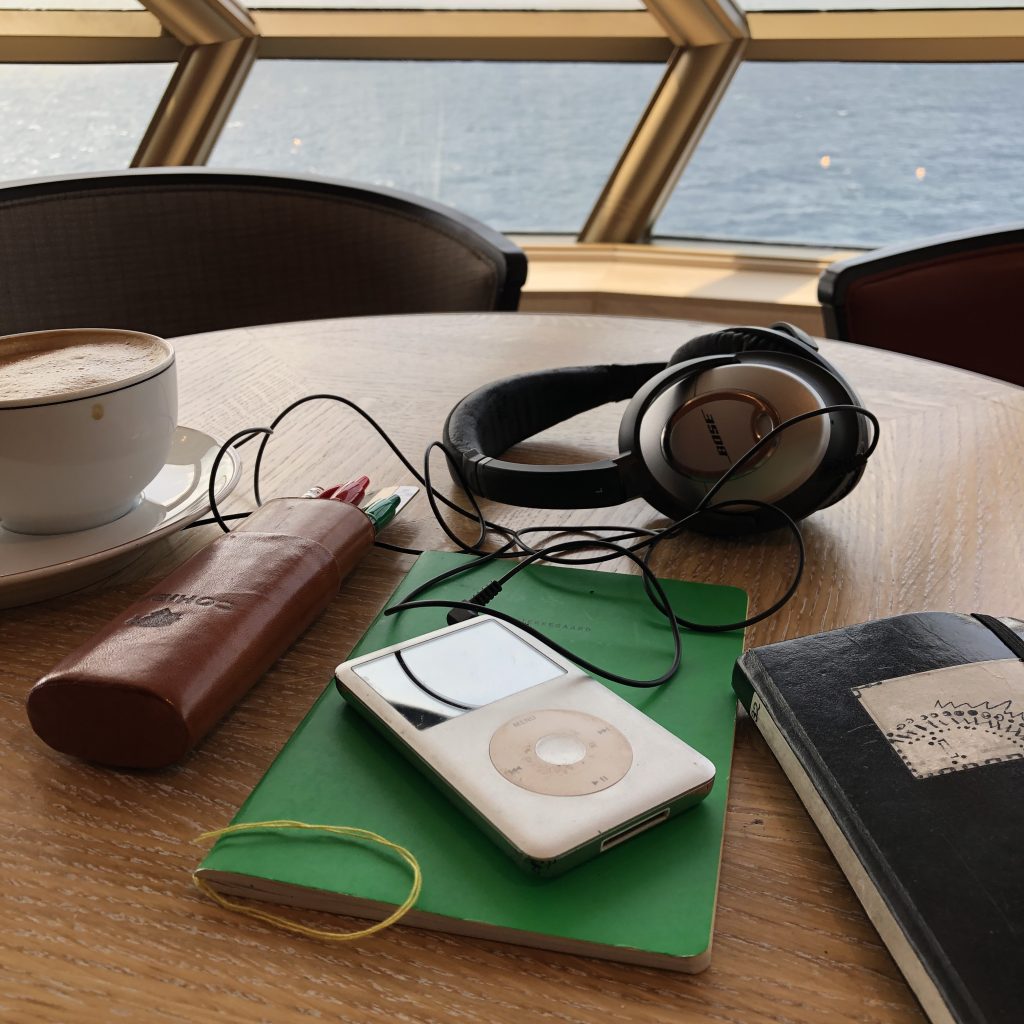 Image: Upon a table sits a green notebook, headphones, pen, and iPhone, by a cup of coffee. Away from the table is an obstructed view of the sea. William Cottman tableau created during a cruise. Photo courtesy of writer.