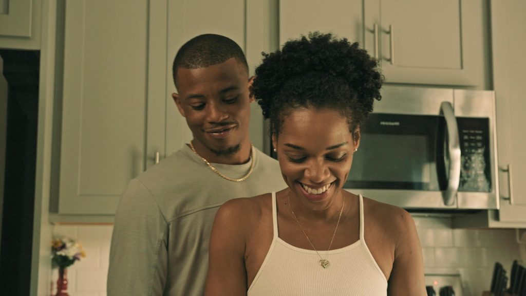 Image: Film still from Red Flags. A Black woman wearing a white tank top looking down and smiling. Behind her is a Black man, looking at the same thing, also smiling. Behind them are grey cabinets and a stainless steel microwave. Photo courtesy of Kierra Payne.