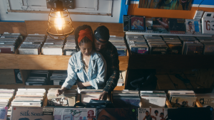 Image: Film still from Tiara West’s “Melanie and Kyle”. A high-angle shot of a Black couple in a bright record store, looking through records. The pair are close together, the man wearing a dark hat and jacket leans over the woman's back. She wears a light jacket and a red bandana. Her right arm is crossed over her stomach. Image courtesy of Tiara West.