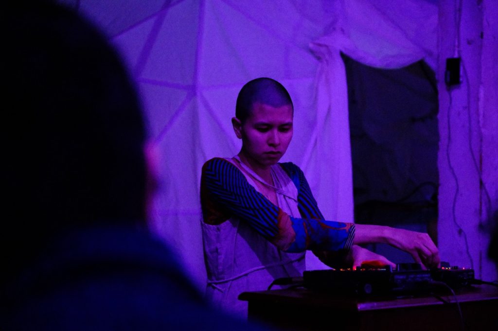 Image: Maya Nyugen performs at EXP/VOX. She is standing in front of a geometric dome and adjusting the settings of a soundboard as she performs. The light on her is purple. She looks intently downward. Photo by Michael Wellvang. Image courtesy of Anna Johnson.