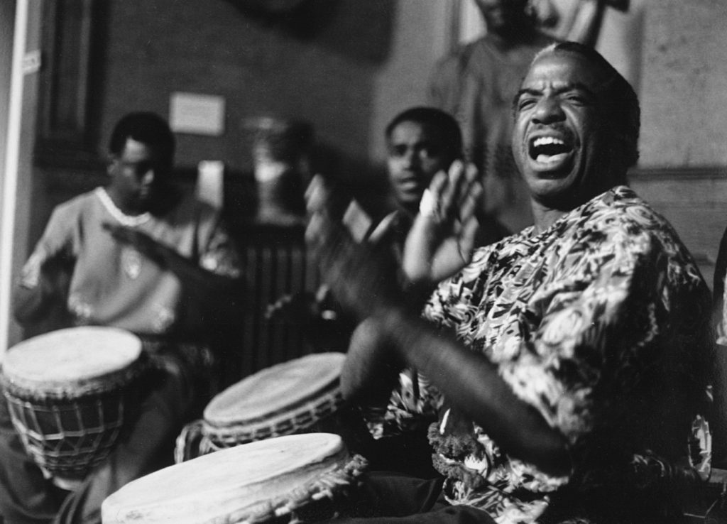 Image: Sylvester "Sunshine" Lee leads a group of young drummers in practice. The photo is in black and white, the trio are caught mid-motion. East St. Louis, 1998. Photo by Theo Pelletier.