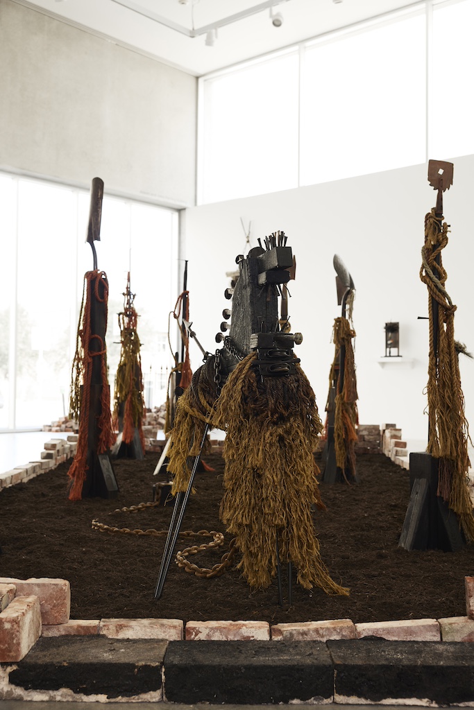 Image: Great Rivers Biennial 2024: Ronald Young, installation view, Contemporary Art Museum St. Louis, September 6, 2024–February 9, 2025. Six tall works made up of textiles, wood, and other media stand in dirt lined with bricks in a gallery. The entire piece is comprised of shades of brown, yellow, and orange. Photo by Izaiah Johnson.