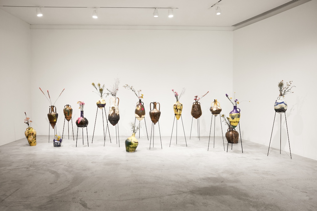 Image: Great Rivers Biennial 2024: Ronald Young, installation view, Contemporary Art Museum St. Louis, September 6, 2024–February 9, 2025. In a gallery space, 12 vessels with flowers sit in stands and four vessels with flowers sit on the gallery floor. Photo by Izaiah Johnson.