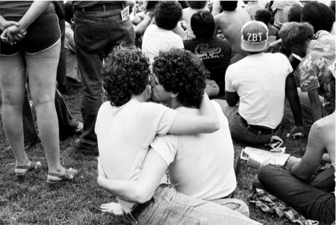 Image: Diana Solís, Couple at Gay Pride Rally, Chicago, Archival Piezographic Print professionally framed with archival glass, 11'' x 16''. A black and white photograph shows a crowd of people sitting outdoors on a grassy area. In the foreground, two individuals with curly hair are sitting close together, facing each other, and kissing. They are both wearing casual clothing, with one person’s arm wrapped around the other’s back. Photo courtesy of Chicago Art Department. 