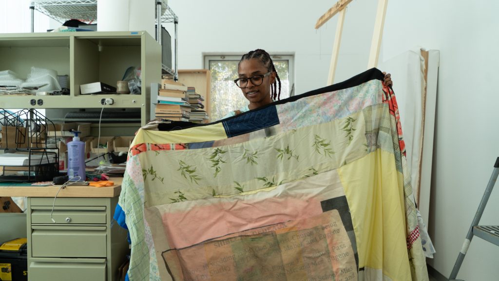 Image: Dedeaux-Norris is pictured holding a patterned quilt which they created. They are pictured center in front of a window. A tool/work shelf with items such as scissors, fabric, paper, tape and markers lay on the shelf. Image by Ian Carstens.