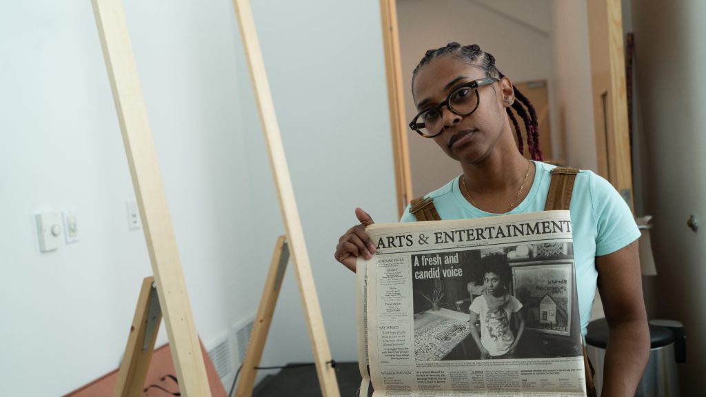 Image: Dedeaux-Norris holds up a newspaper that shows a cover story image of themself early in their career. The article is in the Arts & Entertainment section of a newspaper, the title of the story reads: "A fresh and candid voice," and Dedeaux-Norris is pictured in a scarf and graphic t-shirt surrounded by their work. Norris is holding the newspaper in front of the doorway of their studio. Image by Ian Carstens.