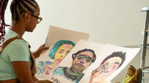 Image: A photo of TJ Norris in her studio holding colorful self-portraits of her various characters. Photo by Ian Carstens.