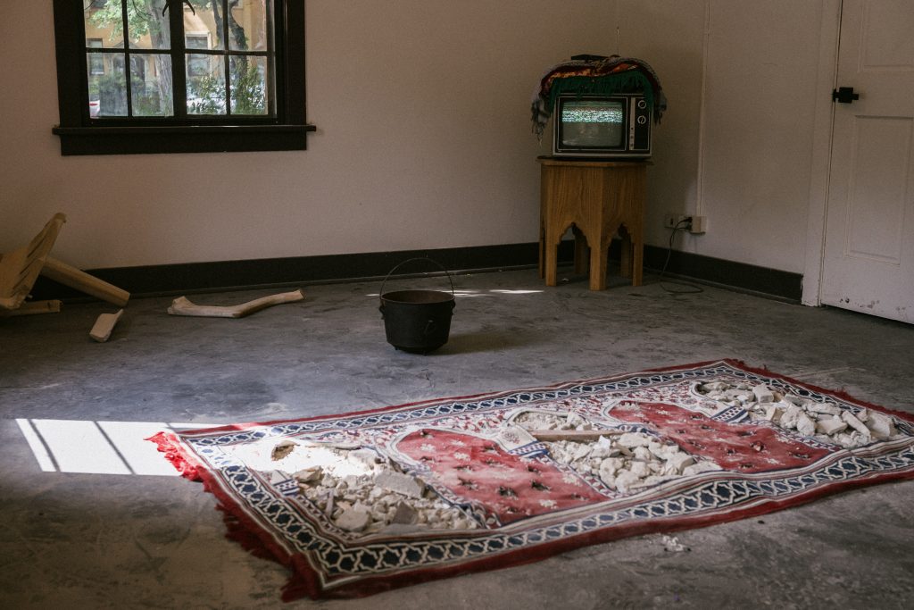 Image: Installation view of At-Atlal. Close up of the eastern wall area with Rug 06 – Rule 38: Intentional targeting of religious buildings during conflict is a war crime under international humanitarian law. A CRT TV displays static in the corner, a cauldron on the ground a few feet in front of it. As of March 2024 Israel has destroyed over 513 Mosques in Gaza is pictured towards the front of the installation image, an uneven rug with rubble strewn across it. Image by Zolt Brown Dunn. Photo courtesy of Comfort Station.