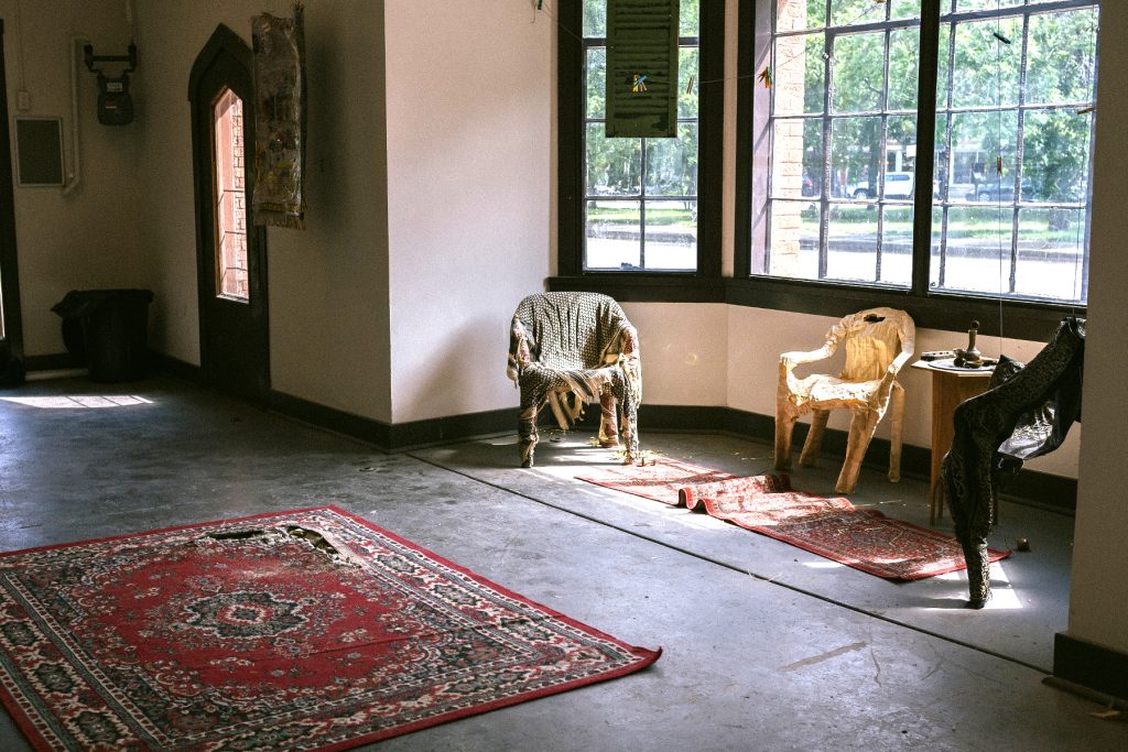 Image: Installation view facing towards the south-faced walls and windows of Comfort Station. Carpet 01: Western Imperialism in the Middle East is pictured on the middle of the floor with works which include Chair 002-4 and Carpet 005 sit in the window-lined enclave of Comfort Station. Rug 05: Self Portrait is seen hanging on the wall behind the enclave. The image space is lit through natural lightning basking through the windows onto the works. Photo by Zolt Brown Dunn. Photo courtesy of Comfort Station.