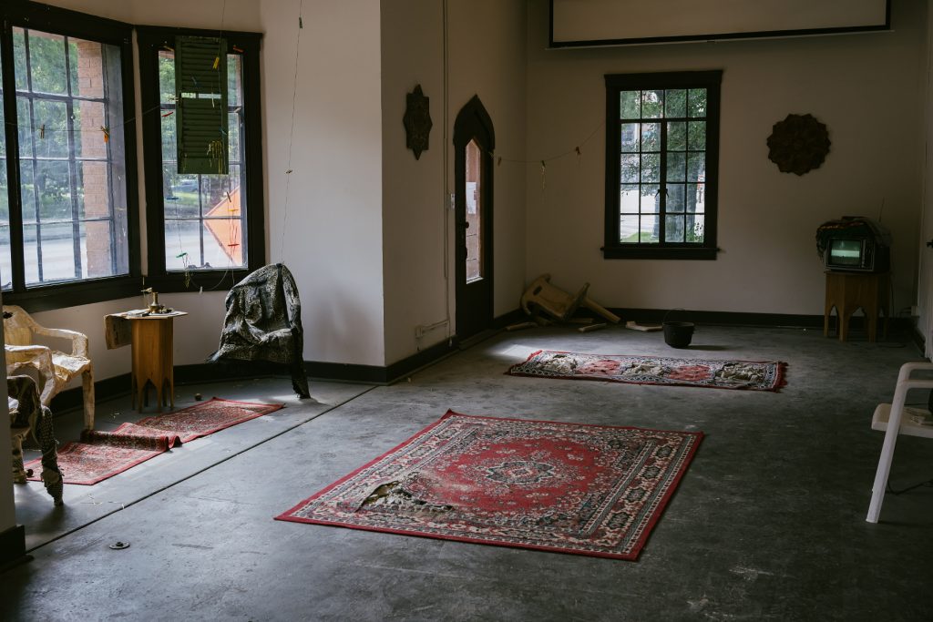 Image: Exhibition view of At-Atlal. The inside of a building with several tall windows looking outside, the light cool on the grey cement floor and several red patterned rugs. Low pieces of furniture are scattered around the room, some destroyed. Photo by Zolt Brown Dunn. Photo courtesy of Comfort Station.