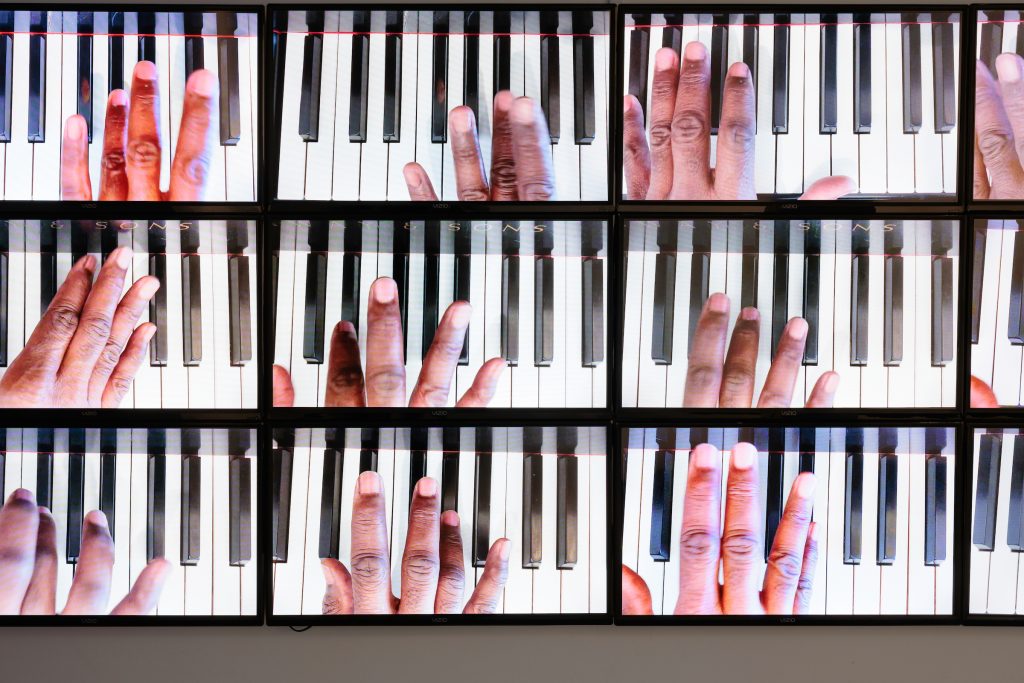 Image: A close up still of Paul Stephen Benjamin's Sonata in Absolute Black All the Black Keys video installation. The still shows twelve tablet-sized monitors showing close up shots of Benjamin's hand playing the black keys on different parts of the piano. Image by Colin Conces, courtesy of Bemis Center.