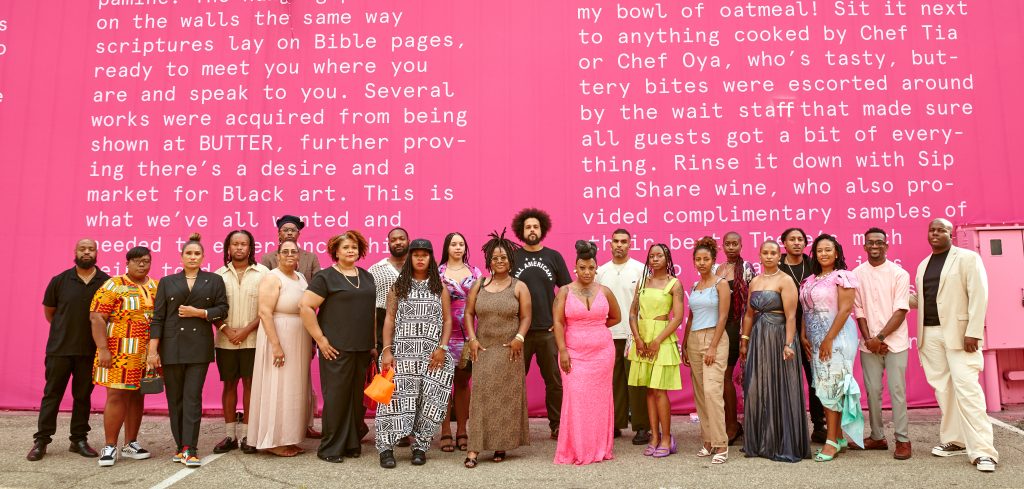 Image: Several of the artists who contributed to BUTTER stand posed in front of the BUTTER branding. The wall behind them is bright pink with white font. Image courtesy of GANGGANG.