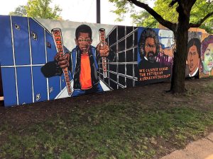 Image: School-to-prison pipeline mural installed at the DuSable Black History Museum and Education Center, created from designs by Juan Luna, Mike Sullivan, Johnny Taylor, Devon Terrell, and Elton Williams. Outside, beyond a small lawna nad tree is a mural of a Black boy near to the foreground wearing an orange shirt with a pipe in either hand, with words on them. Photograph by Sarah Ross, 2021.