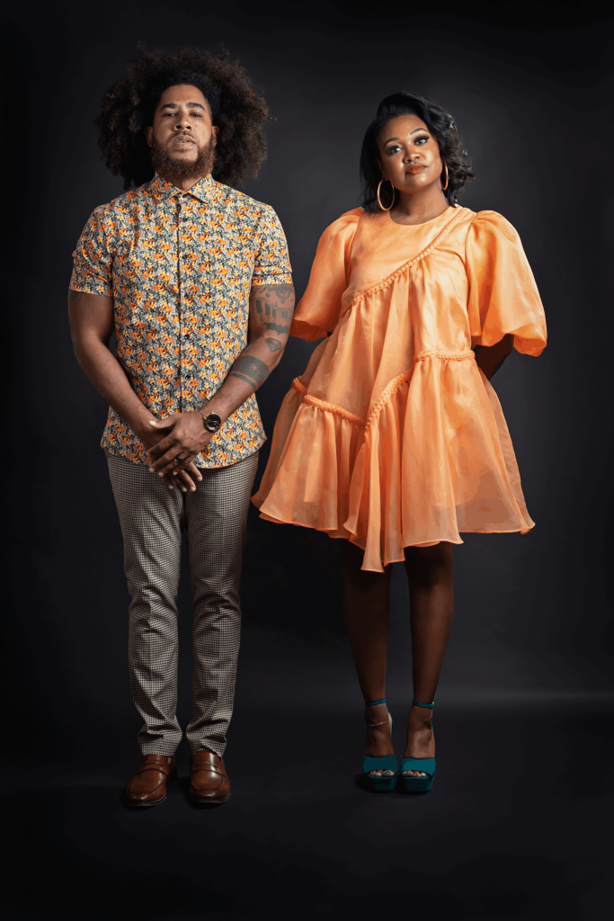 Image: Portrait of Malina Simone and Alan Bacon. Alan Bacon [left] is standing with his arms in front while looking directly at the camera. He is wearing an orange, floral shirt, slacks, and brown shoes. Malina Simone [right] is standing with her hands behind her back with her head slightly tilted while looking directly at the camera. She is wearing a bright orange dress and teal heels. Photographed by Polina Osherov for GANGGANG