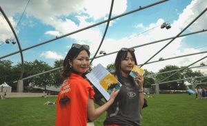 Image: Two people are posed center for the camera under the J. B. Pritzker pavilion holding Urban + Nature Sonic Pavilion brochures. The person posed left is wearing the Urban+Sonic Pavillion orange tee shirt on their back. Behind the two people are pavilion-goers laying on the grass and enjoying the concert, above them is a blue sky. Image by Alex Inglizian, courtesy of Urban+Nature Sonic Pavilion.