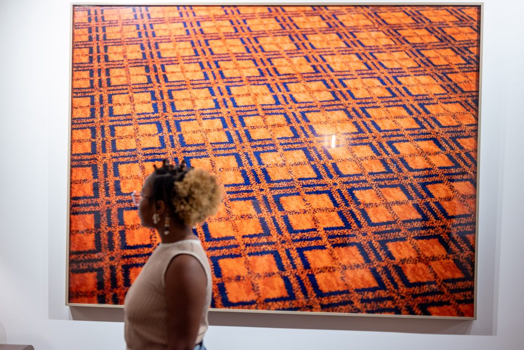 Image: At the Stony Island Arts Bank, a guest of When Clouds Roll Away stands in front of a large photograph of an orange and blue carpet featuring intricate square patterns. September 12, 2024. Photo by Tonal Simmons.