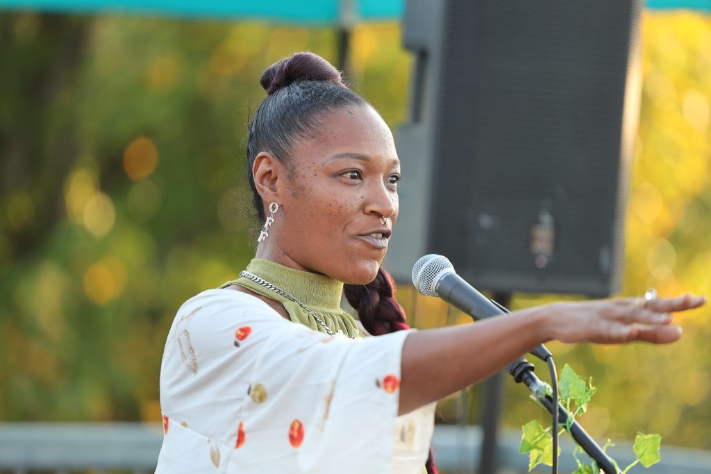 Image: A rooftop open mic at Proof: A Midwest Lit Fest in Indianapolis, IN. The open mic was hosted by That Peace and VOCAB, two popular and long-running Indianapolis open mics. Speaking into the mic is poet Januarie York, host of VOCAB open mic. Photo credit: Indiana Humanities.