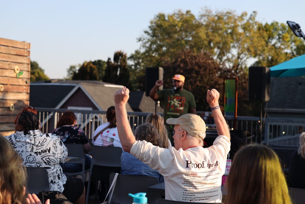 Image: At Proof: A Midwest Lit Fest, a person standing in front of a crowd speaking into a microphone raises his fist. In the foreground, an audience member raises both fists in excitement. Photo credit: Indiana Humanities.