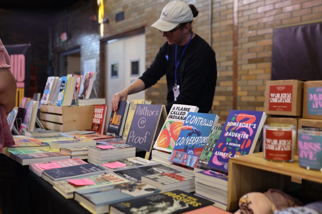 Image: A person standing behind a table of books on display for visitor to peruse at Proof: A Midwest Lit Fest in Indianapolis, IN. Photo credit: Indiana Humanities. 