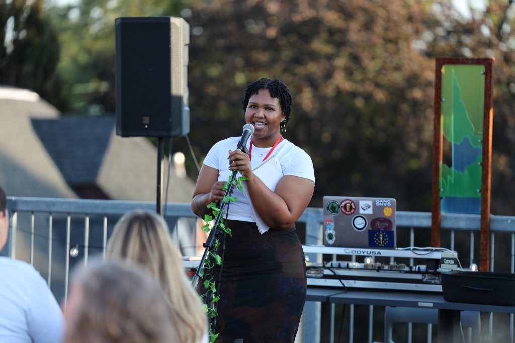 Image: A rooftop open mic at Proof: A Midwest Lit Fest in Indianapolis, IN. The open mic was hosted by That Peace and VOCAB. Speaking into the mic is Mariah Ivey, host and founder of That Peace open mic. Photo credit: Indiana Humanities.