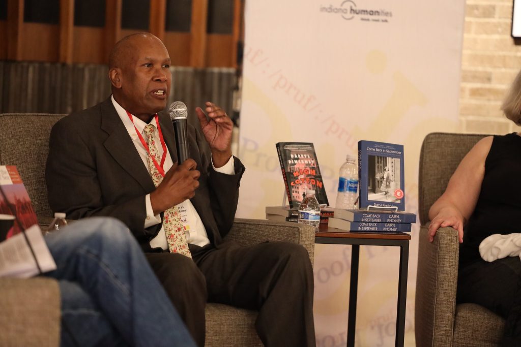 Image: At Proof: A Midwest Lit Fest, novelist and longtime New York Review of Books contributor Darryl Pinckney speaks into a mic during the panel discussion You Can’t Go Home Again. To his right are two of his books: High Cotton: A Novel and Come Back in September: A Literary Education on West Sixty-seventh Street, Manhattan. Photo credit: Indiana Humanities.