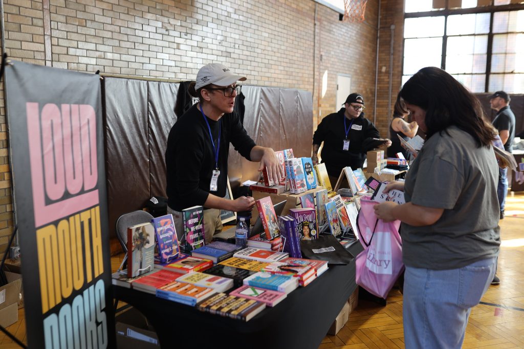 Image: A person standing behind a table of books on display for visitor to peruse at Proof: A Midwest Lit Fest in Indianapolis, IN. The vendor is local bookstore Loudmouth Books. Photo credit: Indiana Humanities. 