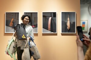 Image: A person holds a backpack while standing in front of a wall of photographs by artist Chang-Ching Casper Su. Photo courtesy of the artist.