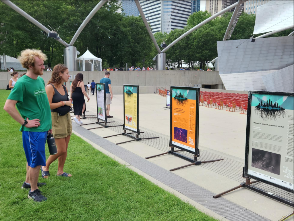 Image: Two people dressed in casual sunny day attire standing in front of a line of UNSP informational posters, each of which describe the work of an individual sound artist, reading the stories behind their pieces. The posters are lined up between the grass and seated area of the pavilion. Image by Alex Inglizian, courtesy of Urban+Nature Sonic Pavilion.