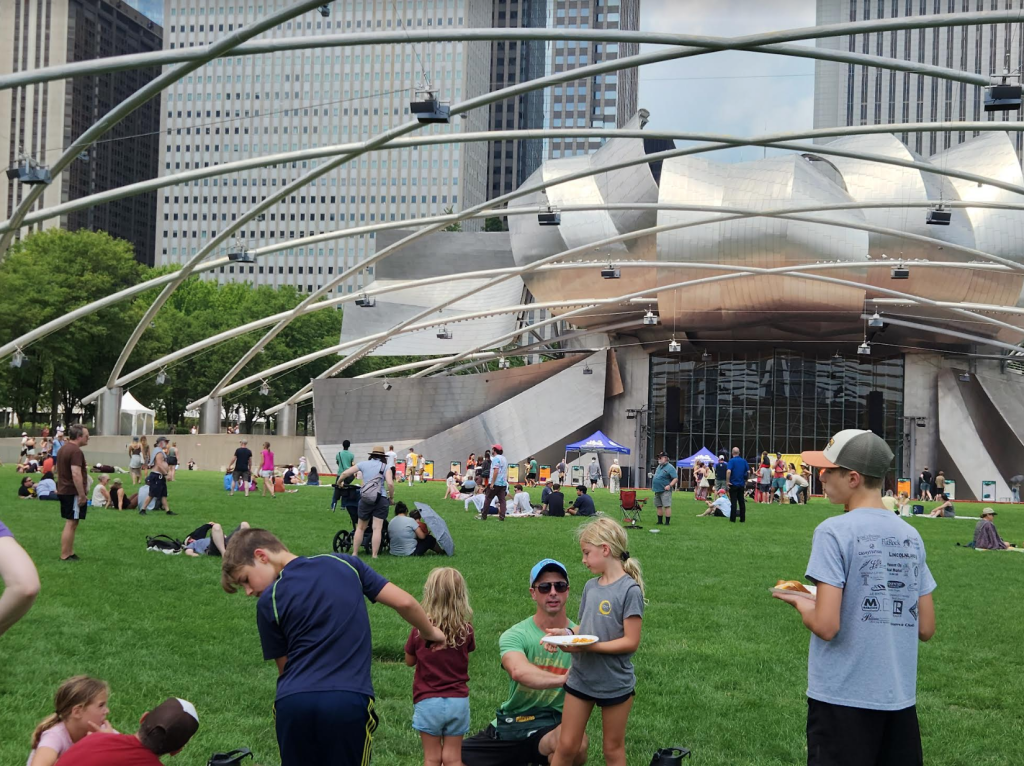 Image: The sound pavilion audience  enjoying the exhibit on a clear, sunny day. A family with children is pictured closest to the camera and there are groups pictured seated and standing throughout the grass. Image by Alex Inglizian.