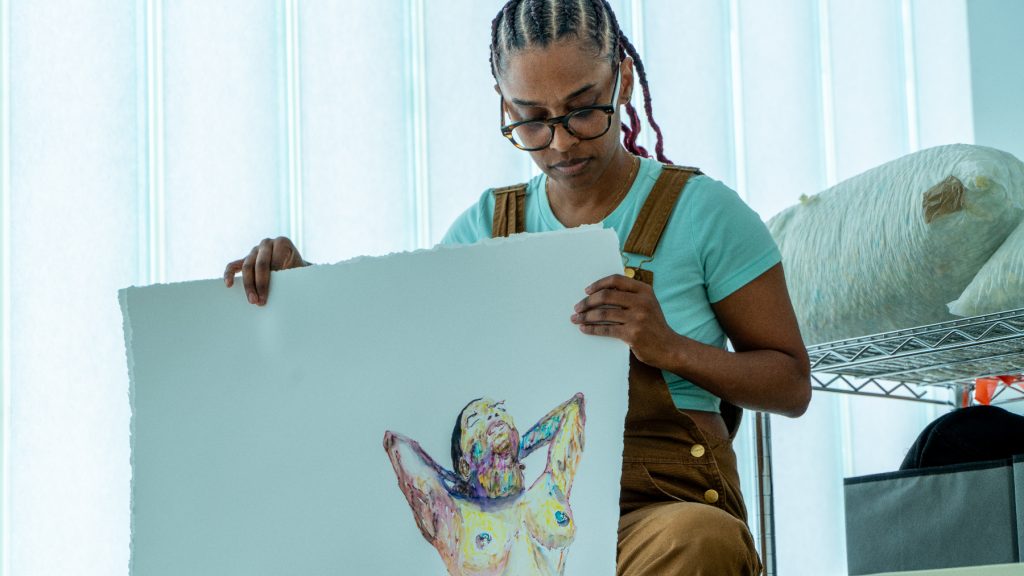 Image: T.J. Dedeaux-Norris is pictured seated on a stool in their studio looking down towards their multi-colored pastel nude in self-portrait. Behind them is a large window and studio materials. In their self-portrait they are pictured leaning back with their head resting on their hands clasped behind their head. The portrait is drawn on white drawing paper. Image by Ian Carstens.