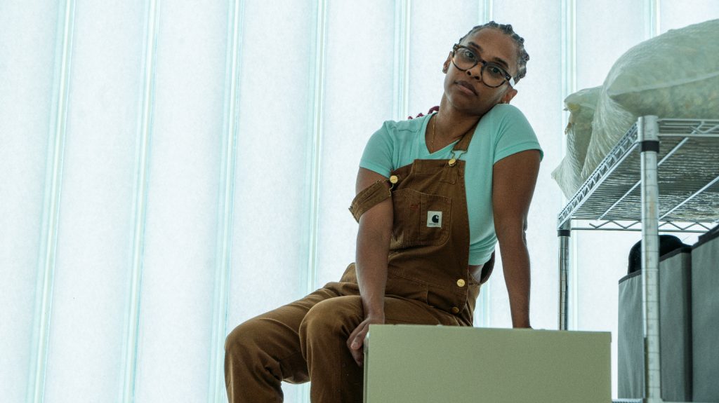 Image: Dedeaux-Norris pictured in their studio seated on a stool near studio materials on metal shelves. Dedeaux-Norris is wearing brown Dickie's overalls, a light blue t-shirt, and large black-rimmed glasses. Their hair is braided into cornrows. Image by Ian Carstens.