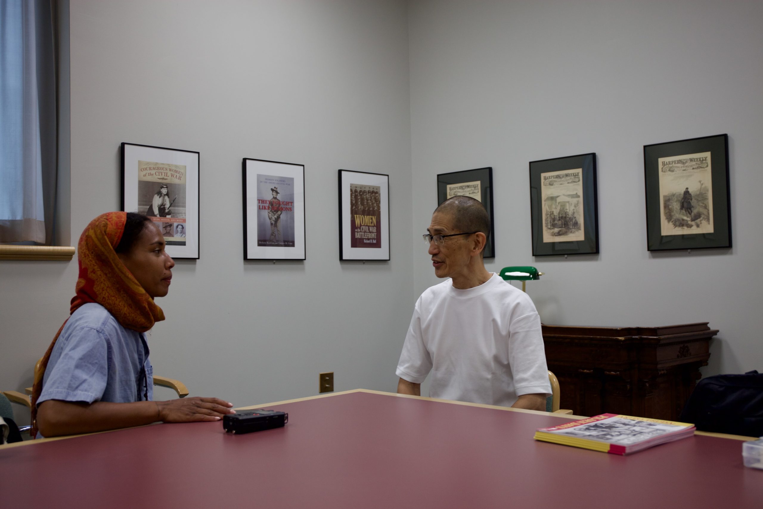 Image: A photograph of Mary Lawson interviewing Akito Tsuda. They sit at a big red table with photographs hung on a wall in the background. Photo by Jose Canas.