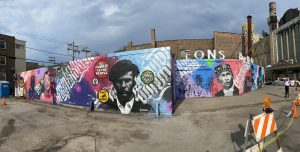 Image: From L-R: Ronnie Malley plays an instrument; an artist for the The Mural Movement stands in front of a work that features Black Panther, Huey P. Newton; Diana Solis holds a camera; Sabba Elahi leads a group in zine making activity at a table in front of a tree mural. Image banner for the Creating Justice Symposium.