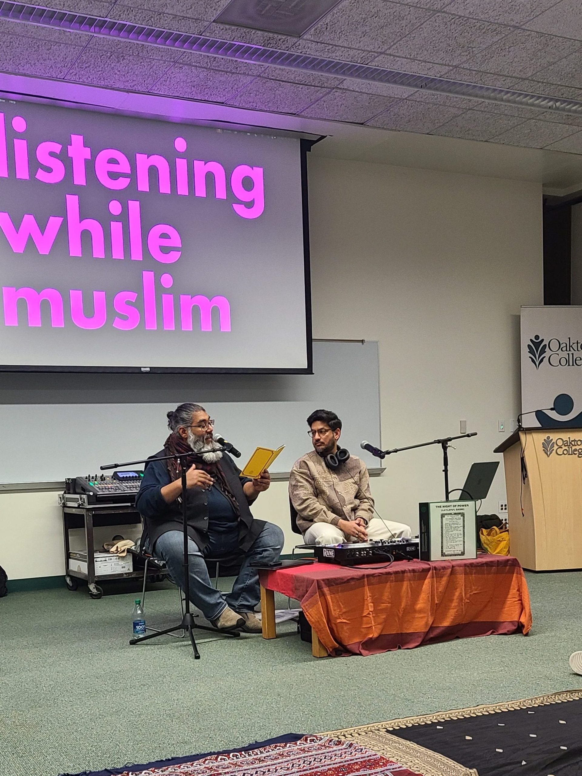 Image: Hosts of the podcast, Listening While Muslim, Asad Ali Jafri (aka DJ Ase Wonder)  and Abdul-Rehman Malik set up chairs, mics and audio recording equipment during a discussion at session three at Oakton College. There are traditional rugs on the floor for the audience to sit on during their conversation.  Image by Chierophant.