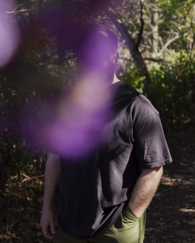 Image: Standing against a forested backdrop, a bearded white person is faced towards the camera.  They wear a black t-shirt with a hand in the pocket of their pants. Their face and part of their torso is obscured by a large out-of-focus purple shape in the foreground. Photo by Joshua Clay Johnson.