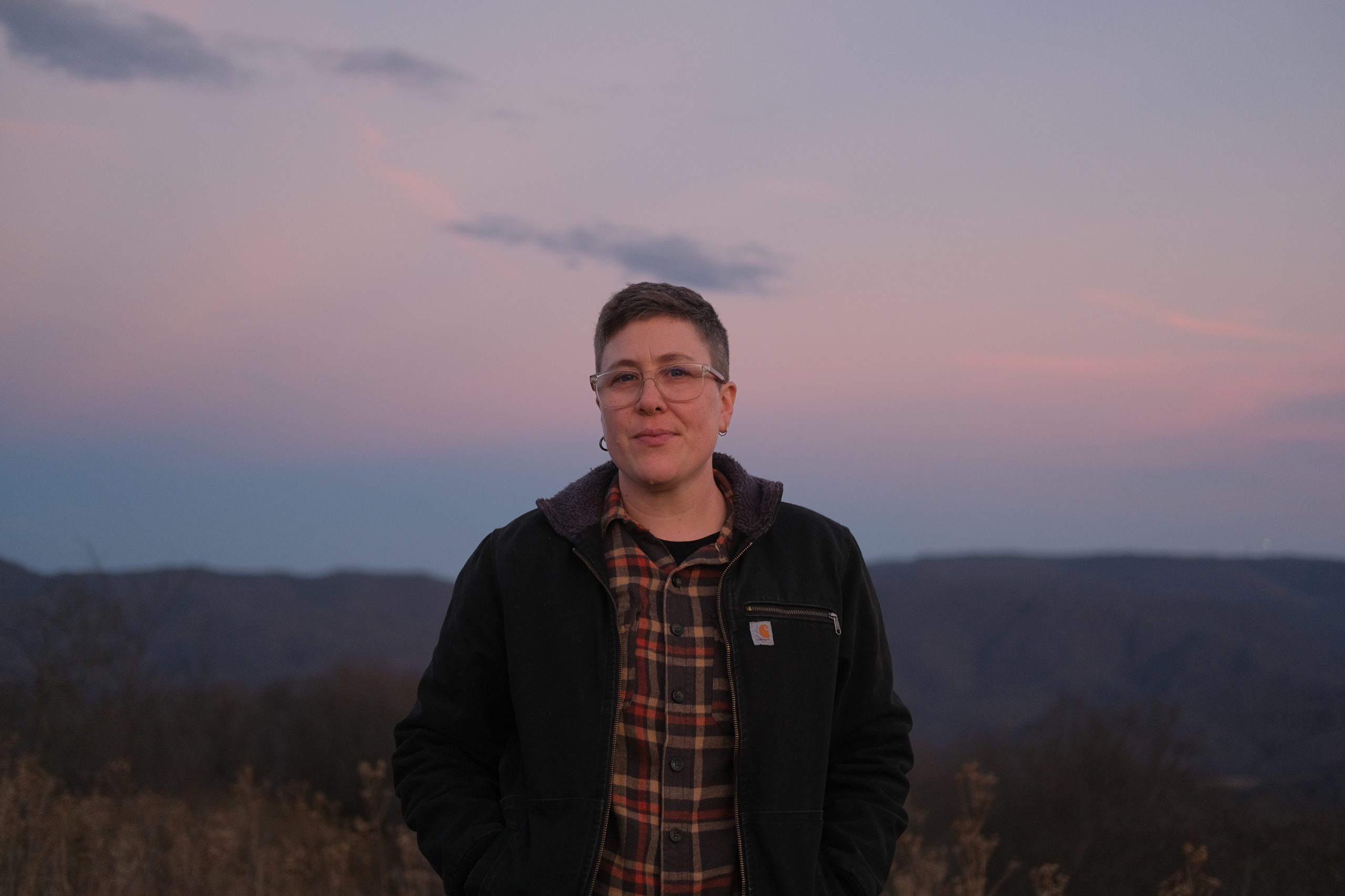 Image: Rae Garringer, wearing a flannel shirt and a dark Carhartt jacket, stands in front of a faded blue and pink mountain skyline in their home of southeastern West Virginia, S’atsoyaha (Yuchi) and Šaawanwaki (Shawnee) lands. Photo by Lou Murrey.