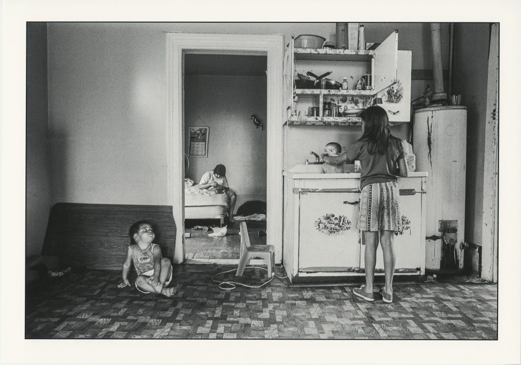 Image: Pilsen, circa 1991-1994, Photographer: Akito Tsuda. Chicago Public Library, Special Collections, Akito Tsuda Photographs, Box 1: Photograph 1.14. In this black and white photograph, a girl washes a baby in the kitchen sink while another child sits on the floor looking at her. In the bedroom in the back, someone who might be the mother sits on the bed sewing a shirt.