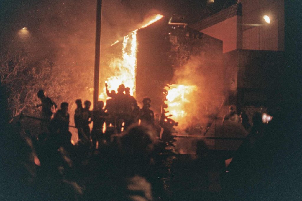 Image: Photo of the 3rd Precinct on fire, 2020. A building can be seen on fire in the background while a group of people are silhouetted in the foreground. Photo sourced from CrimeThinc.com.