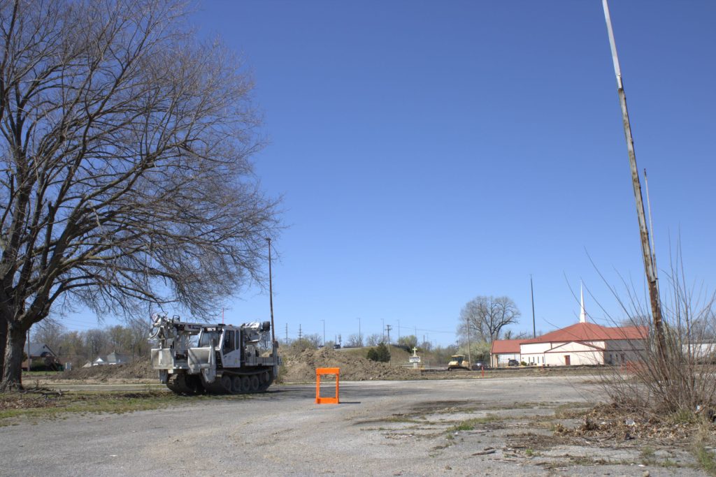 Image: This time the place marker foregrounds the 11th church you will pass traveling west down State street, Greater Saint Mark Church of God in Christ. In the background of the photo on the right is a church, with a construction truck to the left. In the center of the composition is a bright orange garden box. Allena Brazier, "Building Abandonment" series, photography with hand built garden box, East St. Louis, 2022. Photo courtesy of the artist. 