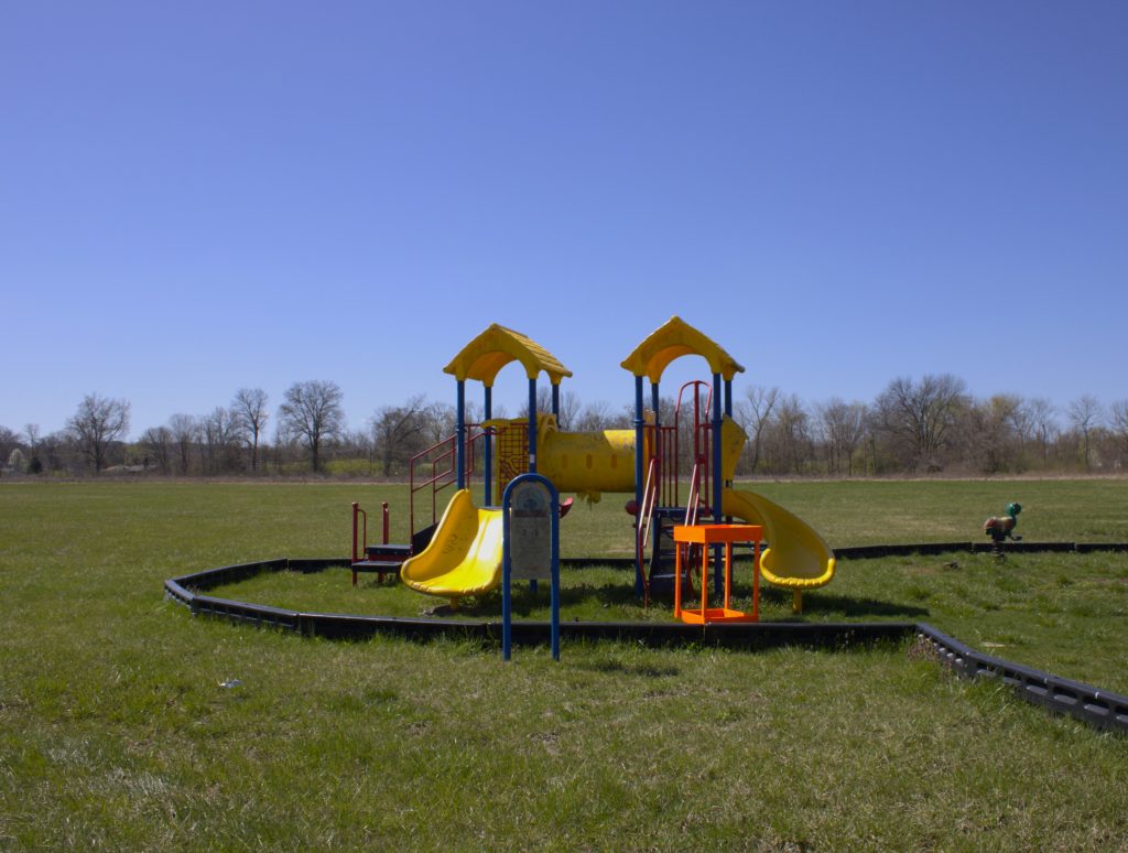 Image: This playground is located at Frank Holten State Park, 4500 Pocket Road, East St. Louis IL 62205. According to Spectrum News: “ . . . Gov. J.B. Pritzker and others announced 55 [electric vehicle] chargers would be placed at state parks and Department of Natural Resource sites. One of the first is already in place at Frank Holten State Park . . .”. The image shows a yellow, blue, and red playground with a line of trees behind it. A bright orange garden box has been placed next to the playground. Allena Brazier, "Building Abandonment" series, photography with hand built garden box, East St. Louis, 2022. Photo courtesy of the artist. 