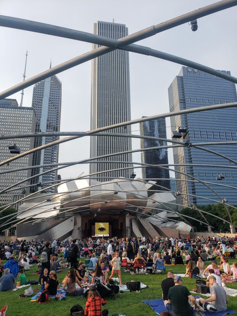 Image: An image of the UBSP during a busy afternoon performance. Grounds of people are pictured seated towards the back of the Pavilion area and people standing close together towards the front of the Pavilion near the stage. It's an overcast day. Image by Alex Inglizian, courtesy of Urban+Nature Sonic Pavilion.