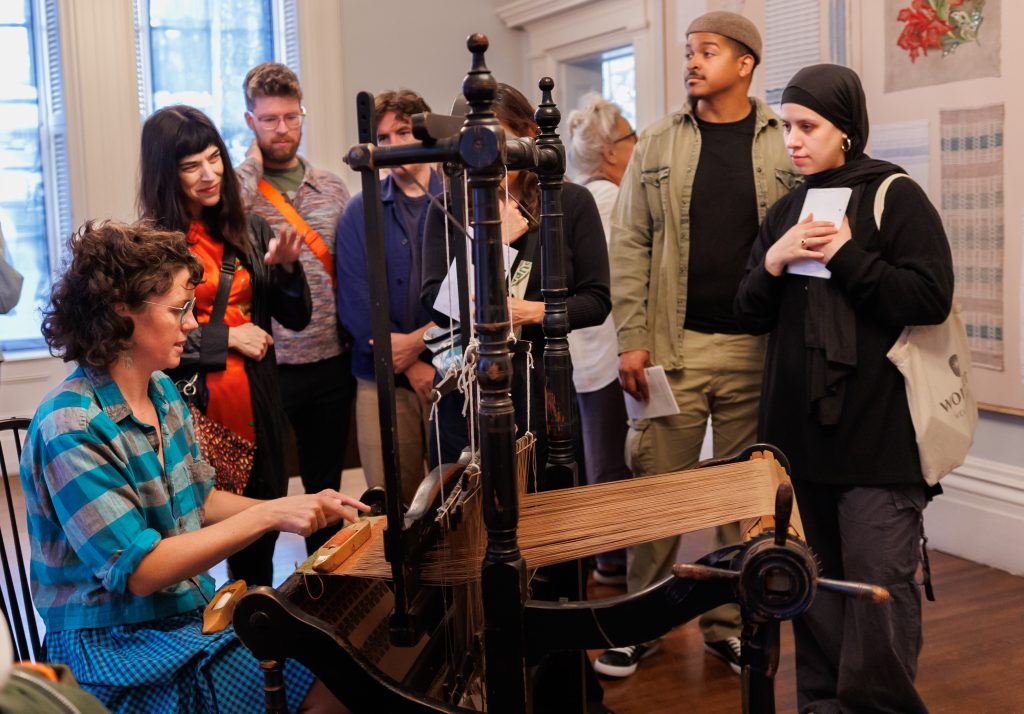 Image: Radical Craft: Arts Education at Hull-House, 1889-1935 opening at the Jane Addams Hull-House Museum. Several people gathered around a loom to watch a demonstration. Courtesy of the Jane Addams Hull-House Museum.