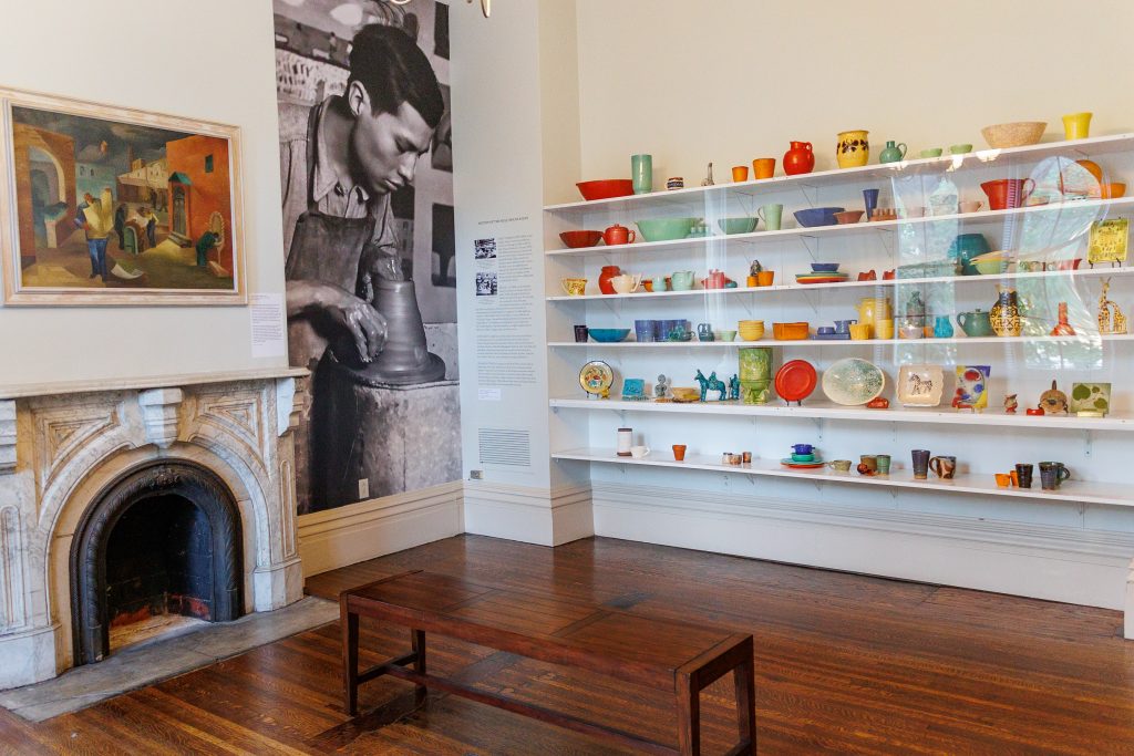 Image: Radical Craft: Arts Education at Hull-House, 1889-1935 opening at the Jane Addams Hull-House Museum. An artwork above a fireplace, phototex on the wall of a person wheel throwing, and six shelves of multicolored ceramics. Courtesy of the Jane Addams Hull-House Museum.