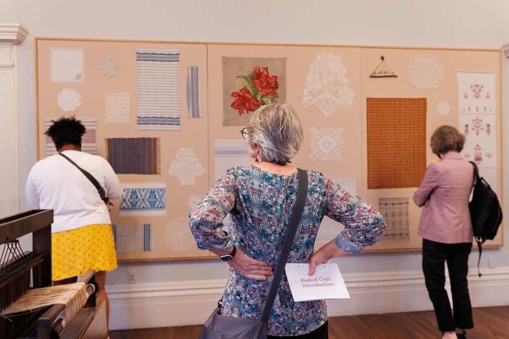 Image: Radical Craft: Arts Education at Hull-House, 1889-1935 opening at the Jane Addams Hull-House Museum. Three people standing in a 'v' looking at a work on the wall with various pattern swatches. Courtesy of the Jane Addams Hull-House Museum.