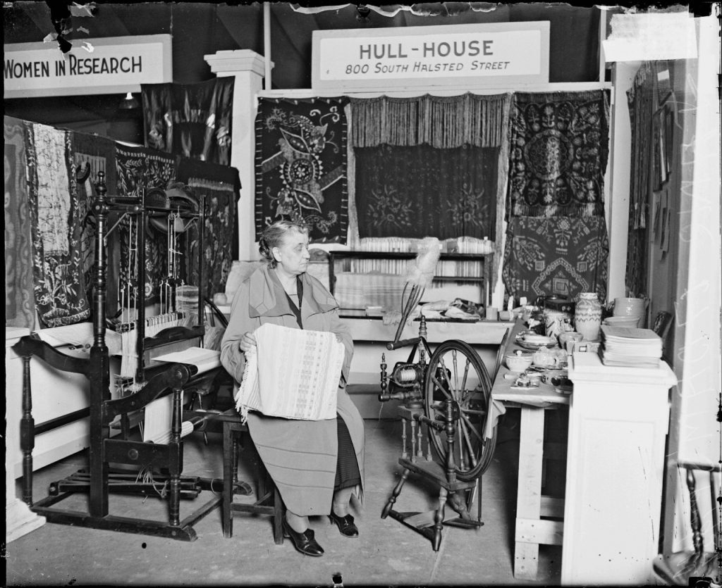 Image: Jane Addams holding a textile between a loom and spinning wheel, possibly at an exhibition of the Labor Museum, Women’s World’s Fair, Chicago, 1927. Full-length portrait of Miss Jane Addams, pacifist, reformer, and founder of the Hull House settlement house, holding a woven garment, sitting next to a spinning wheel and weaving machine in a room in Chicago, Illinois. A sign in the background reads: Hull House, 800 South Halsted Street. A sign in the background on the left side of the image reads: Women in Research. Chicago Daily News Photograph Collection, Chicago History Museum, DN-0084134. 