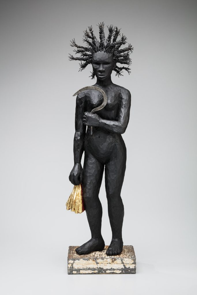 Image: Alison Saar, Topsy and the Golden Fleece, 2017, wood, tar, steel, ceiling tin, wire, acrylic paint, and gold leaf. A nude black figurine sculpture with her hair in a halo of twists stands atop a rustic plinth holding a steel sickle in front of her chest in her left hand and gold in the other. Image courtesy of Toledo Museum of Art.