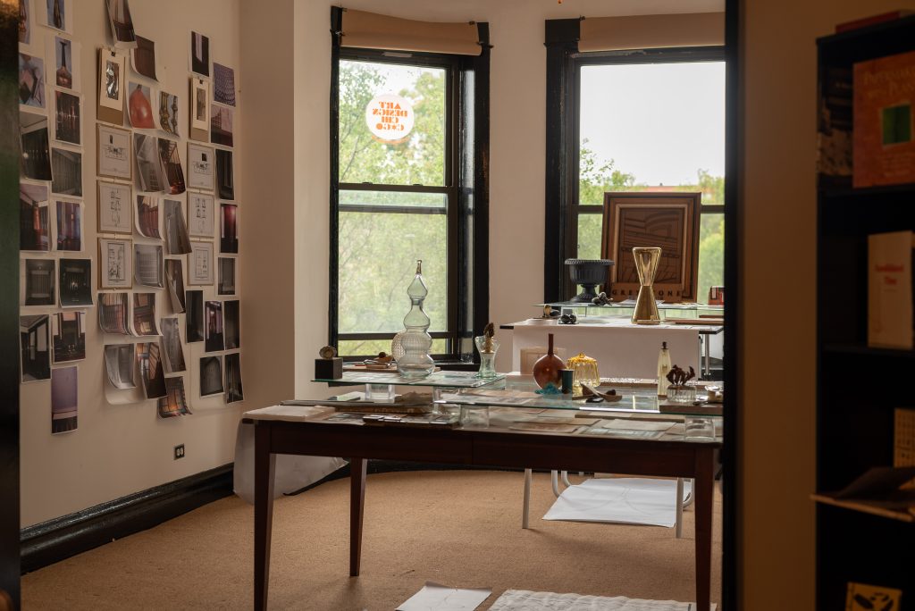 Image: A photograph of an interior room of the Greystone Home and Studio that exhibits printed photographs on the left wall. At the center are two tables that have glass vases, pens, and papers on top. Photograph by Tonal Simmons.