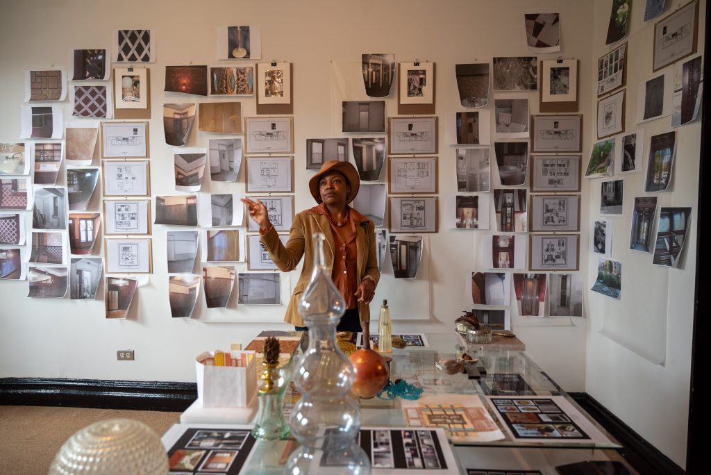 Image: A photograph of Clemenstein Love in her studio standing in front of a wall of printed photographs. In the forefront is a glass desk with various glass vases, photographs, and containers on top. Photograph by Tonal Simmons.