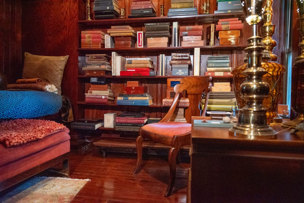 Image: An interior photograph of a library room in the Greystone Home and Studio. A wall of bookshelves holds stacks of hardback books of various colors. On the left of the room is a reading bench and foot stool. On the right of the room is a wood chair and a side table with two gold lamps on them. Photography by Tonal Simmons.